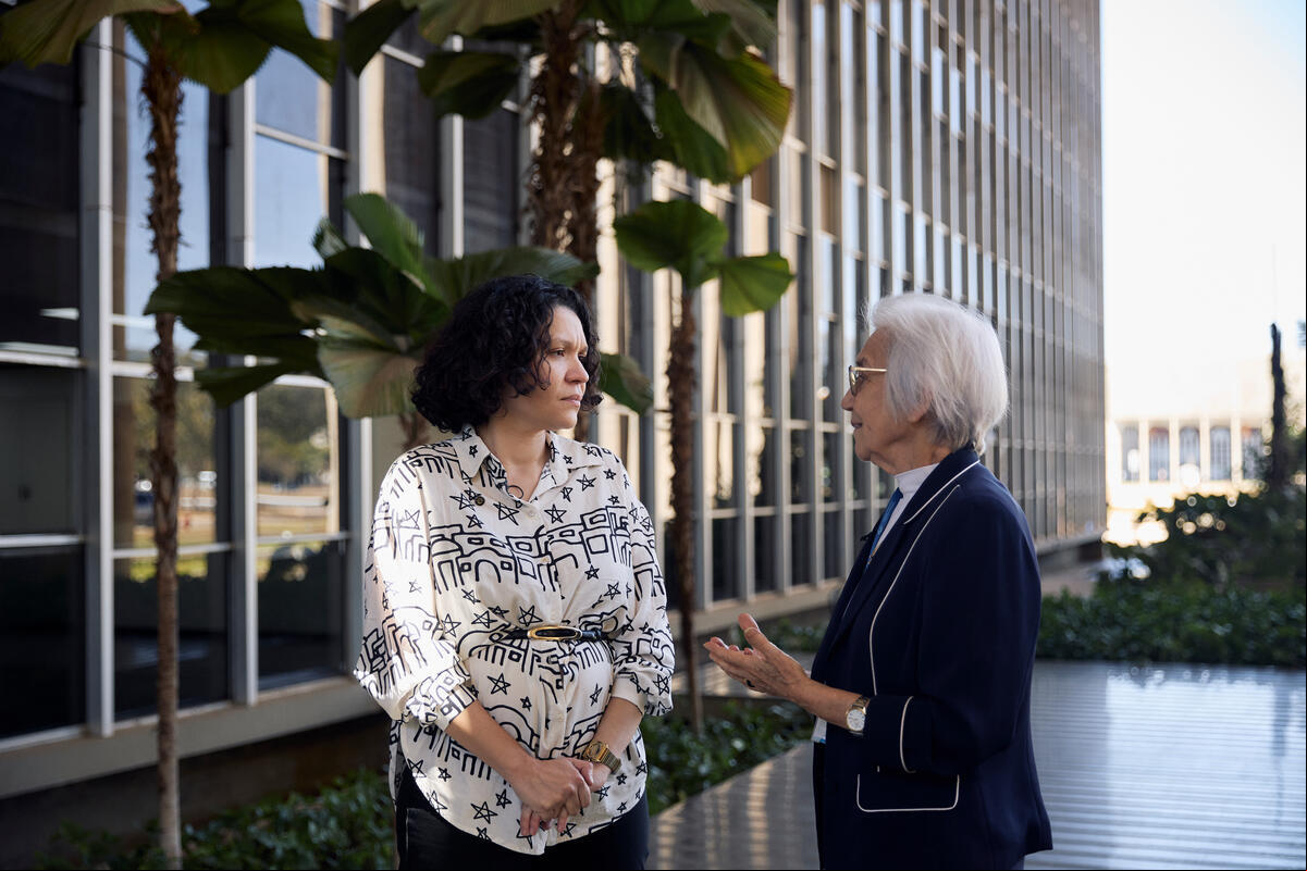 Dos mujeres platican en un pasillo rodeado de plantas en el exterior de un gran edificio institucional