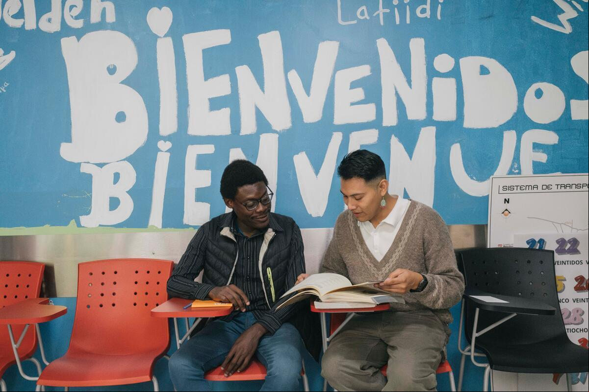 Dos hombres sentados en un salón de clases revisan un libro. Detrás de ellos se lee pintada en la pared la palabra Bienvenidos.