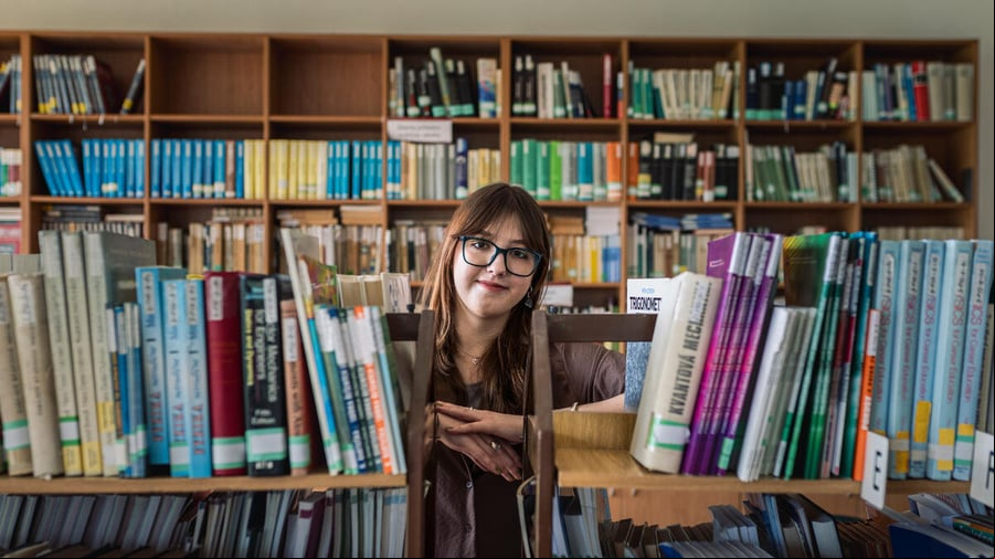 En una biblioteca repleta de libros, detrás de un par de estantes, una joven que usa anteojos y lleva el cabello suelto sonríe a la cámara.